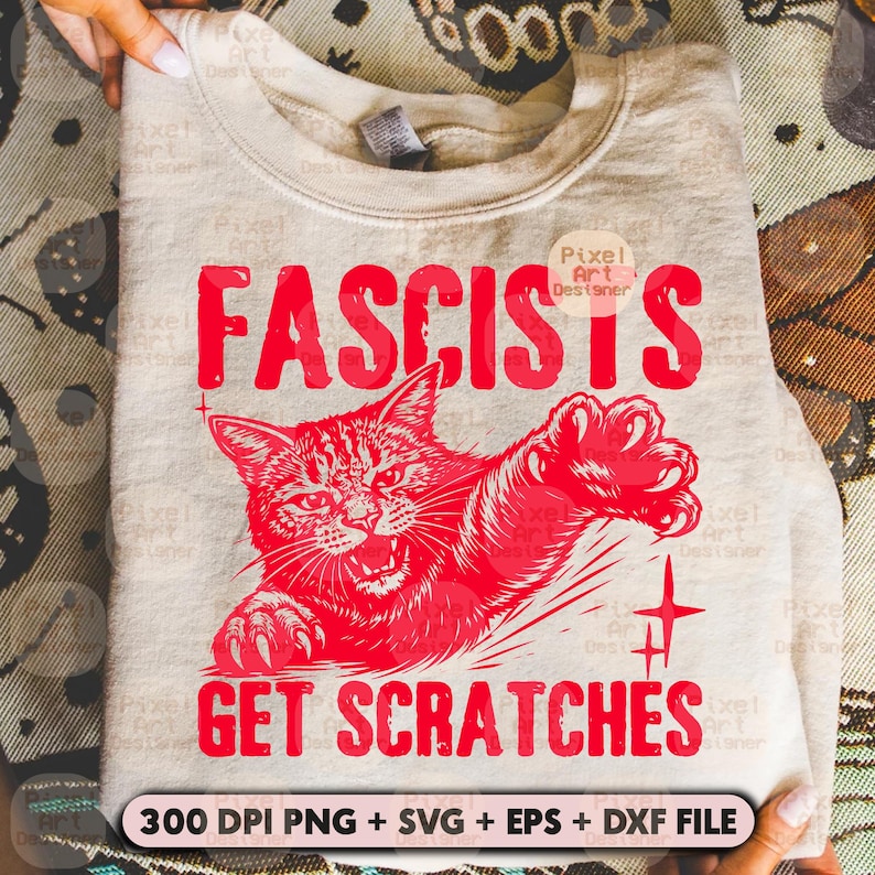 May include: A light beige sweatshirt with the red text "FASCISTS GET SCRATCHES" and a red illustration of an angry cat. The design includes a cross and the text "300 DPI PNG + SVG + EPS + DXF FILE".