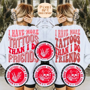 May include: White sweatshirt with red text that reads "I have more tattoos than I do friends". The text is in a retro font style. There are three red circles with black outlines on the sweatshirt. Each circle has a black and white illustration of a skeleton hand with a peace sign and a checkered flag. The text in the circles reads "I have more tattoos than I do friends".