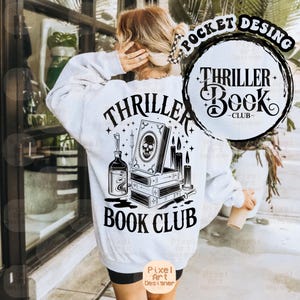 Puede incluir: Sudadera blanca con un diseño gráfico en blanco y negro. El diseño incluye una calavera en un libro, velas y el texto "Thriller Book Club". La sudadera tiene mangas largas.