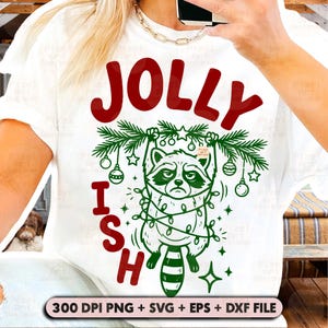 May include: A white t-shirt featuring a graphic of a raccoon tangled in green Christmas lights, hanging from a pine branch. The text JOLLY is printed in red above the raccoon, and the letters I S H are printed in red vertically on the left side.