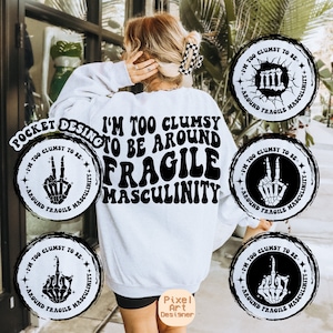 May include: A white sweatshirt with the text "I'm too clumsy to be around fragile masculinity" in black. The text is in a retro font. There are four black and white circular designs with skeleton hands and the text "I'm too clumsy to be around fragile masculinity" around the edge.