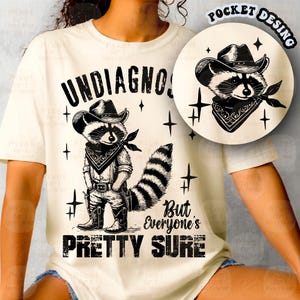 Könnte beinhalten: Ein weißes T-Shirt mit einer schwarz-weißen Illustration eines Waschbären, der einen Cowboyhut und ein Bandana trägt. Der Text "UNDIAGNOSED BUT EVERYONE'S PRETTY SURE" ist auf das Shirt gedruckt.