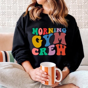 May include: A black sweatshirt with the text "Morning Gym Crew" in colorful retro letters. The person wearing the sweatshirt is sitting on a couch and holding a striped mug.