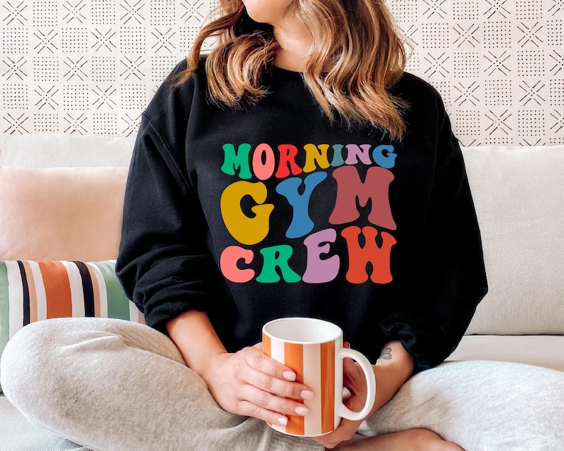 May include: A black sweatshirt with the text "Morning Gym Crew" in colorful retro letters. The person wearing the sweatshirt is sitting on a couch and holding a striped mug.