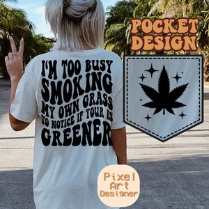 May include: Cream-coloured t-shirt with the text "I'M TOO BUSY SMOKING MY OWN GRASS TO NOTICE IF YOUR IS GREENER" in black, wavy font. A pocket design features a black cannabis leaf and stars. The shirt is a casual wear item.