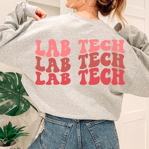 Op de afbeelding: Een grijs sweatshirt met de woorden "Lab Tech" drie keer herhaald in een retro lettertype. De tekst is in tinten roze en rood.