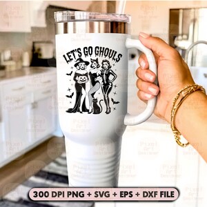 May include: White travel mug with a silver lid and handle. The mug features the text "LET'S GO GHOULS" above a graphic of three women in Halloween costumes, with bats and crosses. A hand holds the mug.