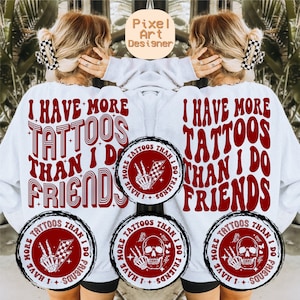 May include: White sweatshirt with red text that reads "I have more tattoos than I do friends". The text is in a retro font style. There are three circular designs on the sweatshirt, each with a skull and crossbones and the text "I have more tattoos than I do friends".