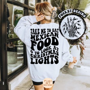 May include: A white sweatshirt with the text "TAKE ME TO EAT MEXICAN FOOD & TO LOOK AT CHRISTMAS LIGHTS" in black. A circular graphic on the right features a skeleton hand holding Christmas lights. The sweatshirt is a casual style.