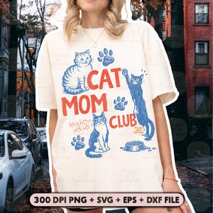May include: Cream-colored t-shirt with a graphic design featuring the text "CAT MOM CLUB" in red and blue, along with illustrations of cats and paw prints. The phrase "life is better with cats" is also included.