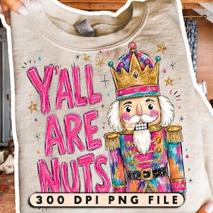 May include: A beige sweatshirt featuring a colorful nutcracker illustration wearing a gold crown and the bright pink text Y'ALL ARE NUTS. The design includes small gold stars and multi-colored dots, with the text 300 DPI PNG FILE at the bottom.