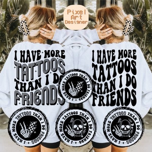 May include: A white sweatshirt with black text that reads "I have more tattoos than I do friends". The text is in a retro font style. There are three black and white circular designs on the sweatshirt, each with a skull and crossbones, a hand with a peace sign, and a checkered flag.