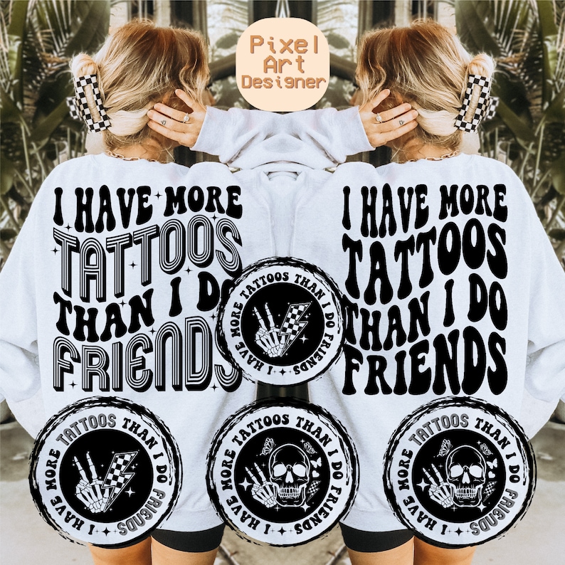 May include: A white sweatshirt with black text that reads "I have more tattoos than I do friends". The text is in a retro font style. There are three black and white circular designs on the sweatshirt, each with a skull and crossbones, a hand with a peace sign, and a checkered flag.