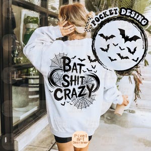 May include: White sweatshirt with a black graphic design on the back that says "BAT SHIT CRAZY" with spiderwebs and bats. A circular design with bats is on the upper right. The sweatshirt has long sleeves.