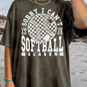 May include: A green t-shirt with a white graphic that says "Sorry I can't it is softball season". The graphic features a softball with a checkered pattern and a heart.