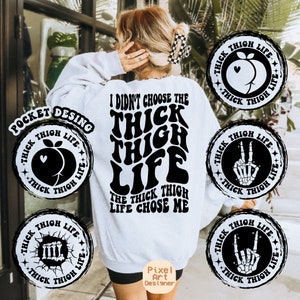 May include: A white sweatshirt with black text that reads "I didn't choose the thick thigh life, the thick thigh life chose me". There are four black and white circular designs around the sweatshirt with the text "Thick Thigh Life" and different graphic designs.