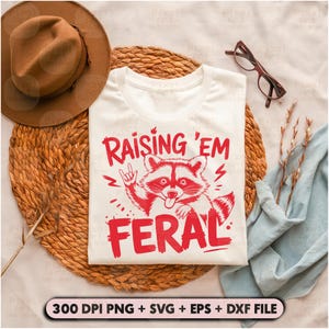 K&ouml;nnte beinhalten: Wei&szlig;es T-Shirt mit einer roten Grafik eines Waschb&auml;ren mit dem Text "Raising 'Em Feral" darauf gedruckt.