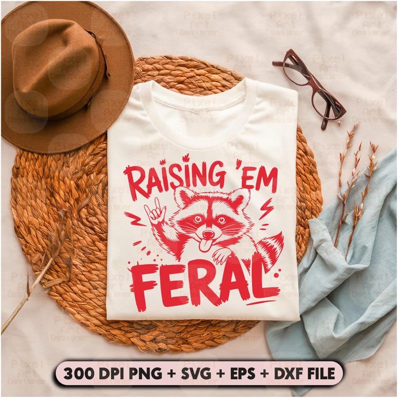 K&ouml;nnte beinhalten: Wei&szlig;es T-Shirt mit einer roten Grafik eines Waschb&auml;ren mit dem Text "Raising 'Em Feral" darauf gedruckt.