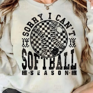 May include: A white sweatshirt with black text that reads "Sorry I Can't It Is Softball Season" with a black and white checkered softball graphic.
