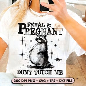 May include: White t-shirt with a black and white graphic of a raccoon and the text "FERAL & PREGNANT DON'T TOUCH ME". The raccoon is standing with its hands in front of it. The shirt also includes the text "300 DPI PNG + SVG + EPS + DXF FILE".