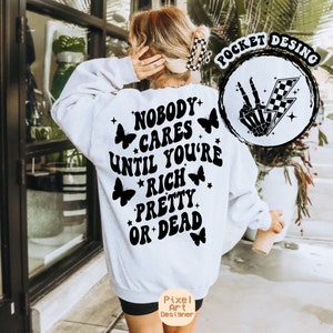 May include: White sweatshirt with black text that reads "Nobody cares until you're rich pretty or dead" with butterfly graphics and stars. The sweatshirt has a pocket on the front with a black and white graphic of a skeleton hand making a peace sign with a lightning bolt and checkered flag.