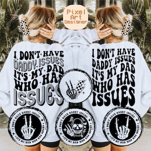 May include: A white sweatshirt with black text that reads "I don't have daddy issues it's my dad who has issues". The text is in a retro font style. There are three black and white illustrations of a skeleton hand with a peace sign, a skull, and a skeleton hand with a rock and roll hand gesture.