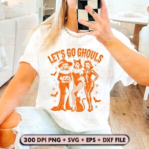 May include: White t-shirt featuring the orange text "LET'S GO GHOULS" and a graphic of three women in Halloween costumes. The design includes a witch, a cat woman, and a skeleton. A black cat and bats are also featured.