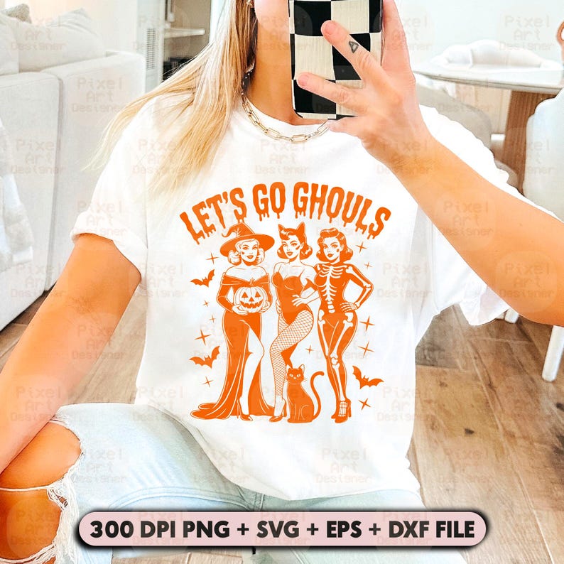 May include: White t-shirt featuring the orange text "LET'S GO GHOULS" and a graphic of three women in Halloween costumes. The design includes a witch, a cat woman, and a skeleton. A black cat and bats are also featured.