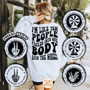 May include: White sweatshirt with the text "I'M LIKE TEN PEOPLE PACKED INTO ONE BODY SPIN THE WHEEL" in black. The design includes circular graphics with the same text and a peace sign hand skeleton. The sweatshirt is a casual style.