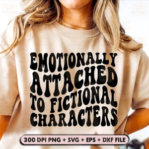 Pode incluir: Uma camiseta bege com texto preto que diz "Emocionalmente ligado a personagens fictícios" em um estilo de fonte retrô. O texto está centralizado na camiseta.
