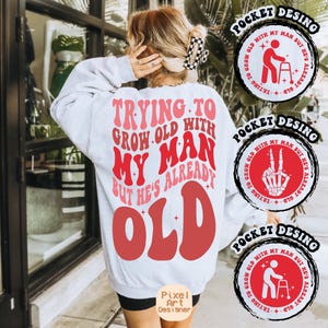 May include: White sweatshirt with a red and pink graphic that says "Trying to grow old with my man but he's already old". The graphic is in a retro font style. There are three pocket designs on the image, each with a different graphic. The pocket designs are in black and red.