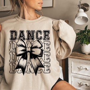 May include: A beige sweatshirt with a black graphic design that says "DANCE" with a large black bow in the center. The graphic is distressed and has a vintage look.