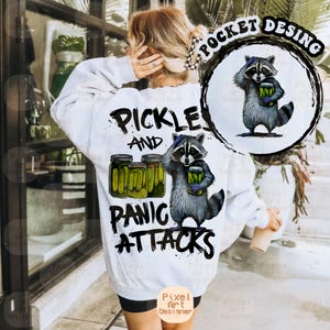 May include: White sweatshirt with a graphic design featuring a raccoon holding a jar of pickles, with the text "Pickle and Panic Attacks". The design includes three jars of pickles and a smaller raccoon graphic on the upper right.