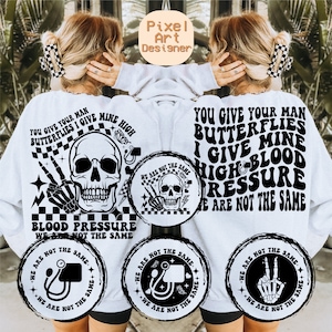 May include: White sweatshirt with black text that reads "You give your man butterflies I give mine high blood pressure we are not the same". The text is surrounded by black and white checkered patterns, skulls, and peace signs.