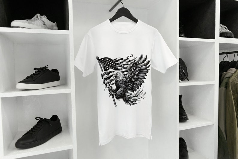 May include: White t-shirt featuring a detailed black and white graphic of an American bald eagle holding a flag. The eagle's wings are spread, and the flag is waving. The shirt is hanging on a black hanger.