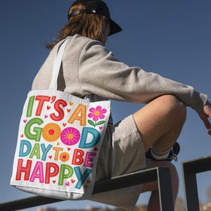 May include: A white tote bag with the text "IT'S A GOOD DAY TO BE HAPPY" in colorful, playful lettering. The bag features a flower graphic and heart accents. The person is wearing a gray sweatshirt, shorts, and a black cap.
