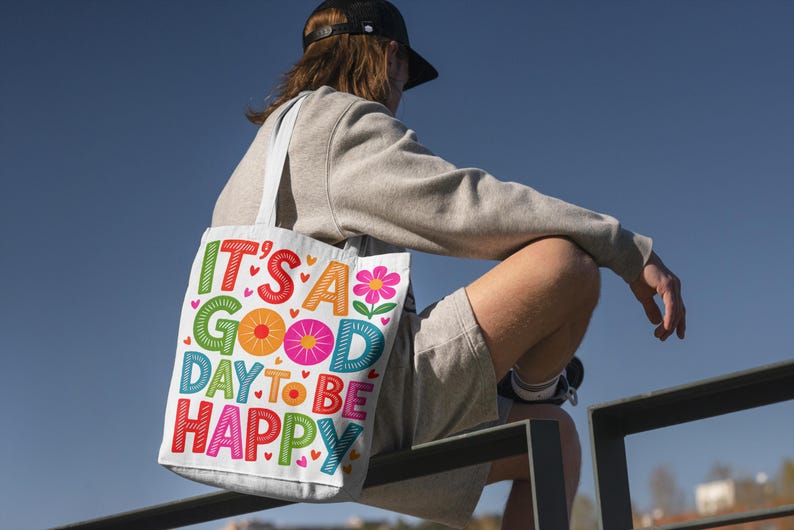 May include: A white tote bag with the text "IT'S A GOOD DAY TO BE HAPPY" in colorful, playful lettering. The bag features a flower graphic and heart accents. The person is wearing a gray sweatshirt, shorts, and a black cap.