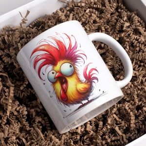 May include: White ceramic mug featuring a cartoon rooster with vibrant red, yellow, and orange feathers and an exaggerated expression. The mug is inside a white box filled with brown crinkle paper.