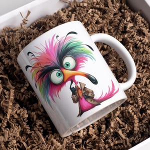 Puede incluir: Taza de cer&aacute;mica blanca con un dise&ntilde;o de p&aacute;jaro de dibujos animados colorido. El p&aacute;jaro tiene grandes ojos verdes, un largo pico negro y un cuerpo rosa. Lleva un abrigo marr&oacute;n y sostiene una peque&ntilde;a bolsa. La taza est&aacute; en una caja blanca llena de papel marr&oacute;n.