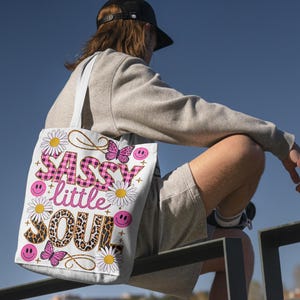 May include: A white tote bag with a pink and black design. The bag features the text "Sassy little Soul" with a pink and black checkered pattern, leopard print, and butterflies.