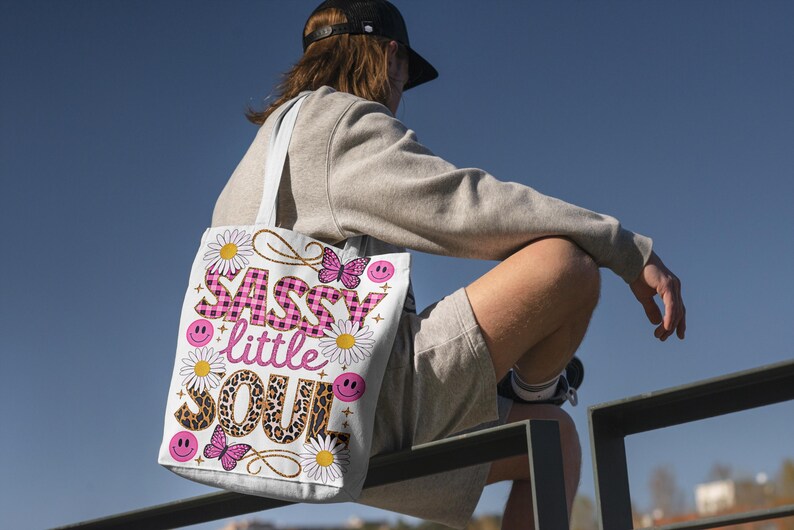 May include: A white tote bag with a pink and black design. The bag features the text "Sassy little Soul" with a pink and black checkered pattern, leopard print, and butterflies.