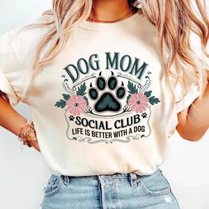 May include: A white t-shirt with a vintage-style graphic that says "Dog Mom Social Club Life is Better with a Dog". The graphic features a black paw print with pink flowers.