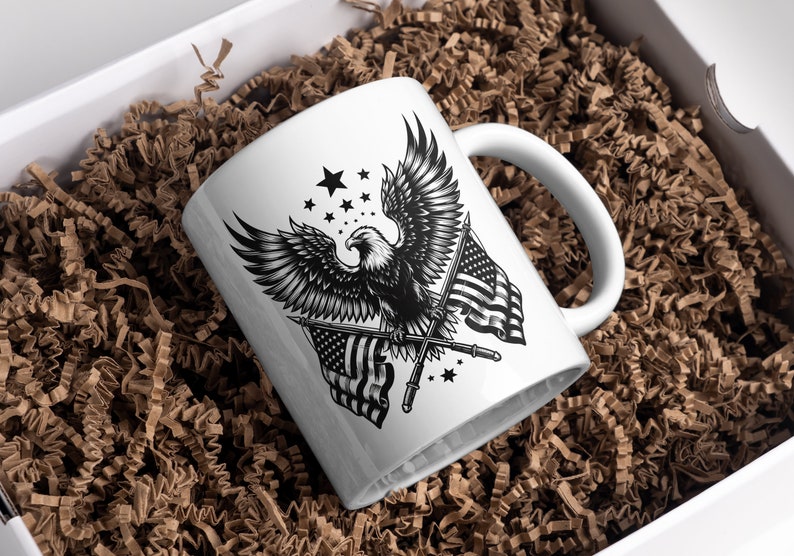 May include: White ceramic mug featuring a detailed black and white illustration of an eagle with outstretched wings, American flags, and stars. The mug is presented in a white box filled with brown crinkle paper.