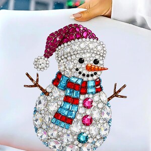 Puede incluir: Un dise&ntilde;o decorativo de mu&ntilde;eco de nieve con un gorro rojo y blanco, una bufanda azul y roja y una nariz de zanahoria. El mu&ntilde;eco de nieve est&aacute; adornado con cristales transparentes y rosas y tiene brazos de ramitas. El dise&ntilde;o est&aacute; sobre un fondo blanco.