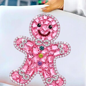 Puede incluir: Un dise&ntilde;o de hombre de jengibre rosa con una apariencia brillante y similar a una gema. La figura tiene una cara sonriente, una pajarita y est&aacute; delineada con cristales transparentes. El dise&ntilde;o se establece sobre un fondo blanco, lo que sugiere un tema navide&ntilde;o.