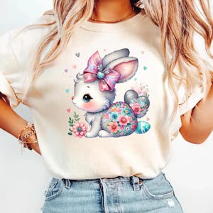 Op de afbeelding: Een wit T-shirt met een kleurrijk cartoonkonijn dat een roze strik draagt en een paasei vasthoudt. Het konijn wordt omringd door bloemen en harten.