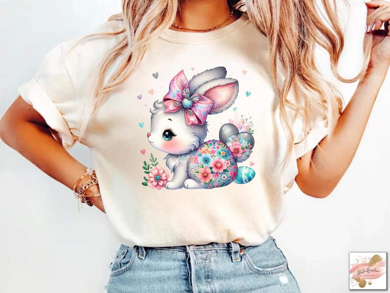 Op de afbeelding: Een wit T-shirt met een kleurrijk cartoonkonijn dat een roze strik draagt en een paasei vasthoudt. Het konijn wordt omringd door bloemen en harten.