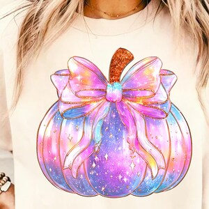 Puede incluir: Camiseta de color claro con una ilustraci&oacute;n en acuarela de una calabaza adornada con un gran lazo colorido. La calabaza es una mezcla de azules, morados y rosas, con detalles de purpurina dorada. El lazo es un degradado de rosas y morados.