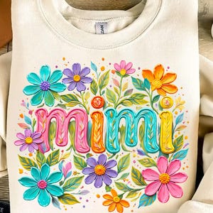 K&ouml;nnte beinhalten: Cremefarbenes Sweatshirt mit dem Wort "Mimi" in bunten 3D-Buchstaben, umgeben von leuchtenden Blumen in Rosa, Blau, Lila und Orange. Gr&uuml;ne Bl&auml;tter und kleine gelbe Akzente vervollst&auml;ndigen das florale Design. Ein gem&uuml;tliches und fr&ouml;hliches Kleidungsst&uuml;ck.