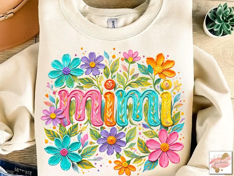 K&ouml;nnte beinhalten: Cremefarbenes Sweatshirt mit dem Wort "Mimi" in bunten 3D-Buchstaben, umgeben von leuchtenden Blumen in Rosa, Blau, Lila und Orange. Gr&uuml;ne Bl&auml;tter und kleine gelbe Akzente vervollst&auml;ndigen das florale Design. Ein gem&uuml;tliches und fr&ouml;hliches Kleidungsst&uuml;ck.